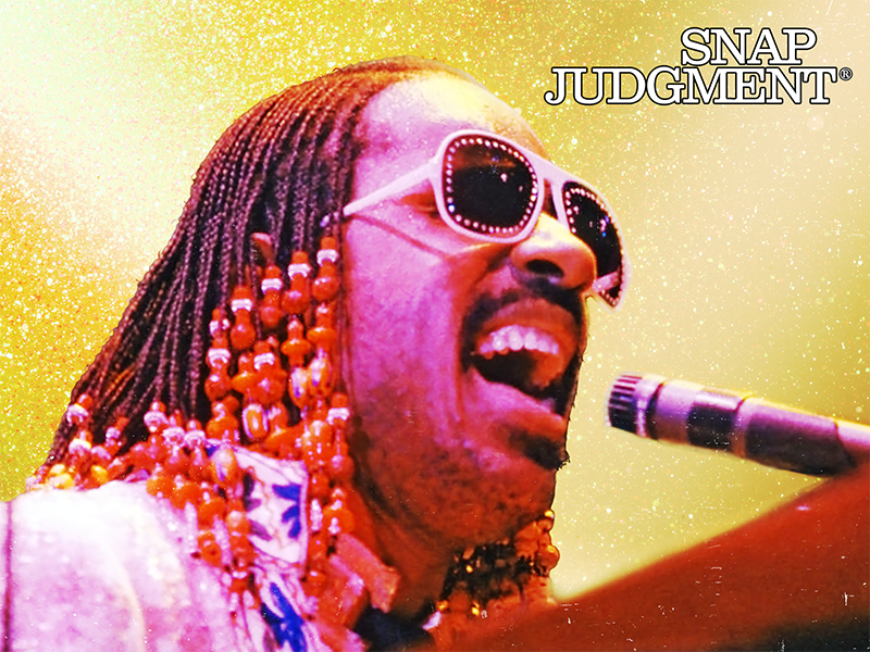 Stevie Wonder is singing into a microphone. He is wearing shades and has braids with colorful beads.
